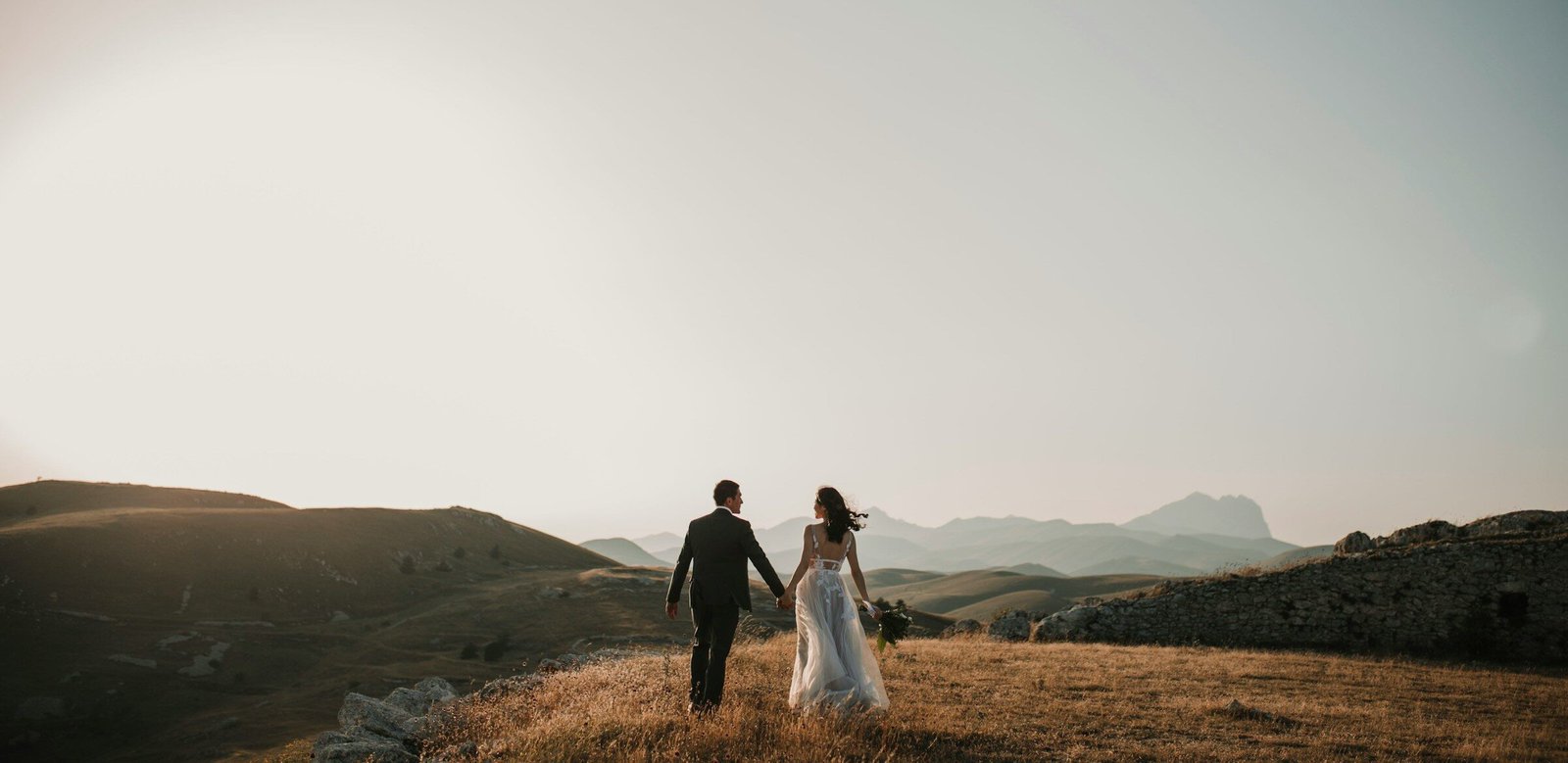 Bride and groom walking hand in hand across a golden hillside at sunset, with mountains in the background.