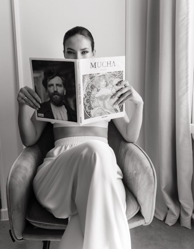 Black-and-white portrait of a woman seated in a chair, reading an art book titled "Mucha."