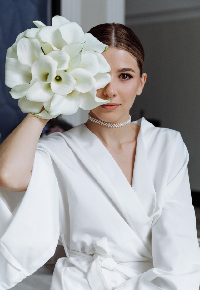 Modern bride wearing a white robe, playfully covering one eye with a calla lily bouquet.
