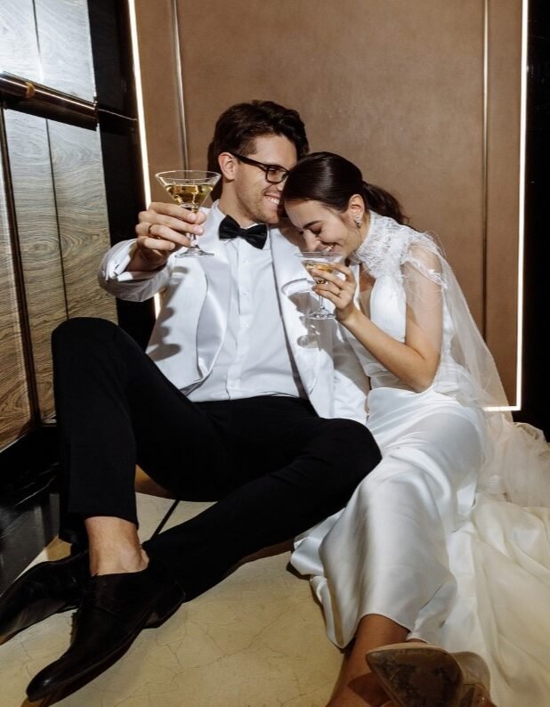 Bride and groom sharing a celebratory champagne toast, seated on the floor in wedding attire.