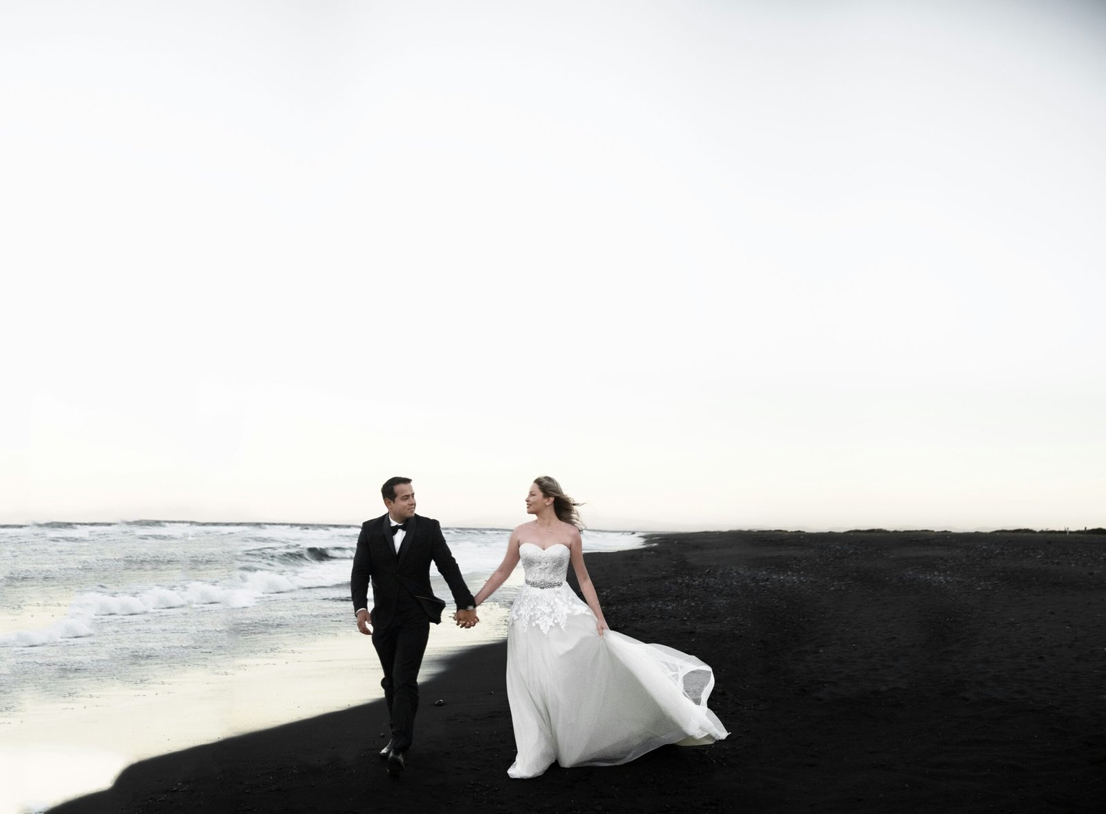 Bride and groom walking hand in hand along a black sand beach, with gentle waves in the background.