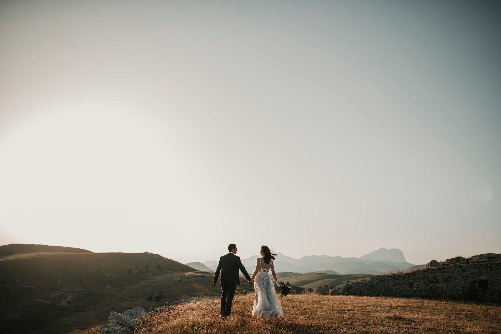 Bride and groom walking hand in hand across a scenic hillside, capturing a romantic moment at sunset.