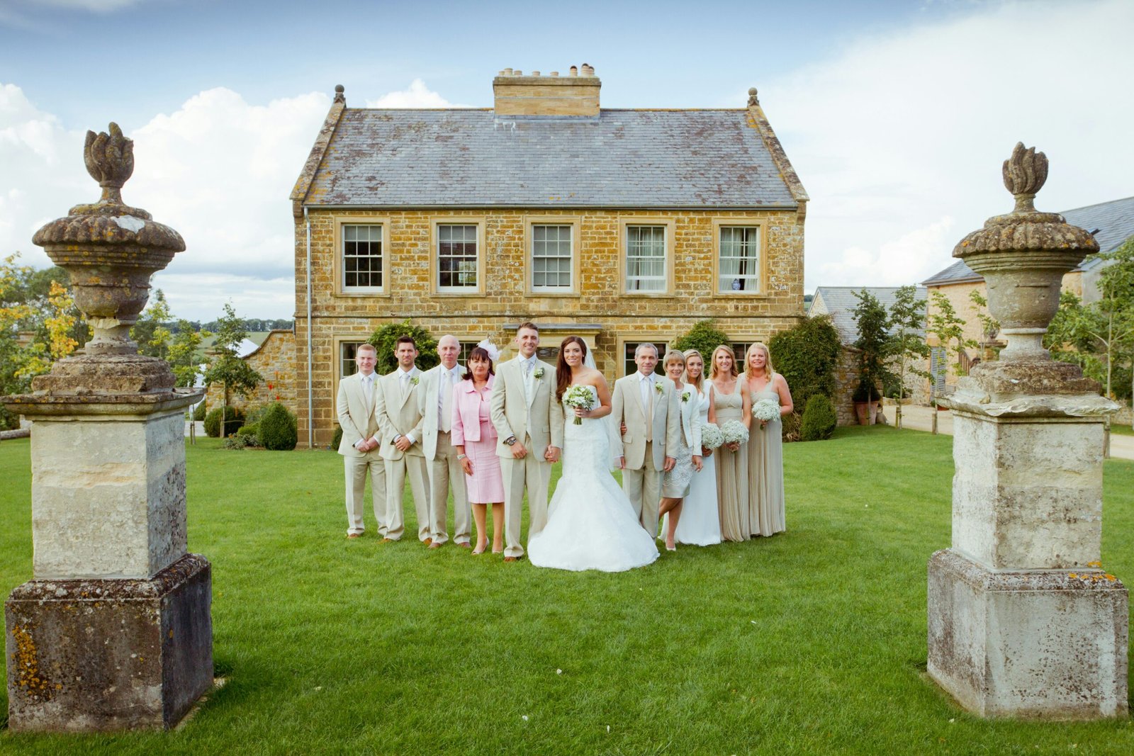 Bride and groom standing with their wedding party in matching cream outfits, posed in front of a rustic stone house.