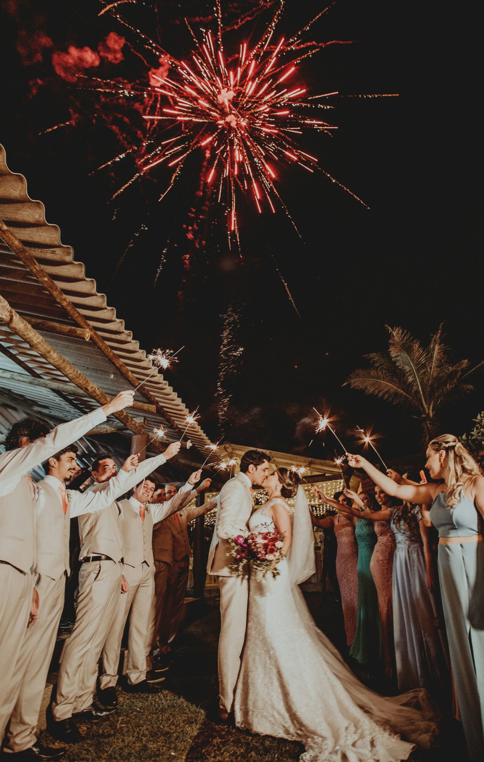 Bride and groom share a romantic kiss under a dazzling fireworks display, surrounded by wedding guests with sparklers.