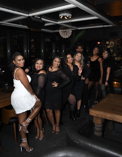 Group of women in stylish black outfits (one in white) smiling and posing at a lively night event.